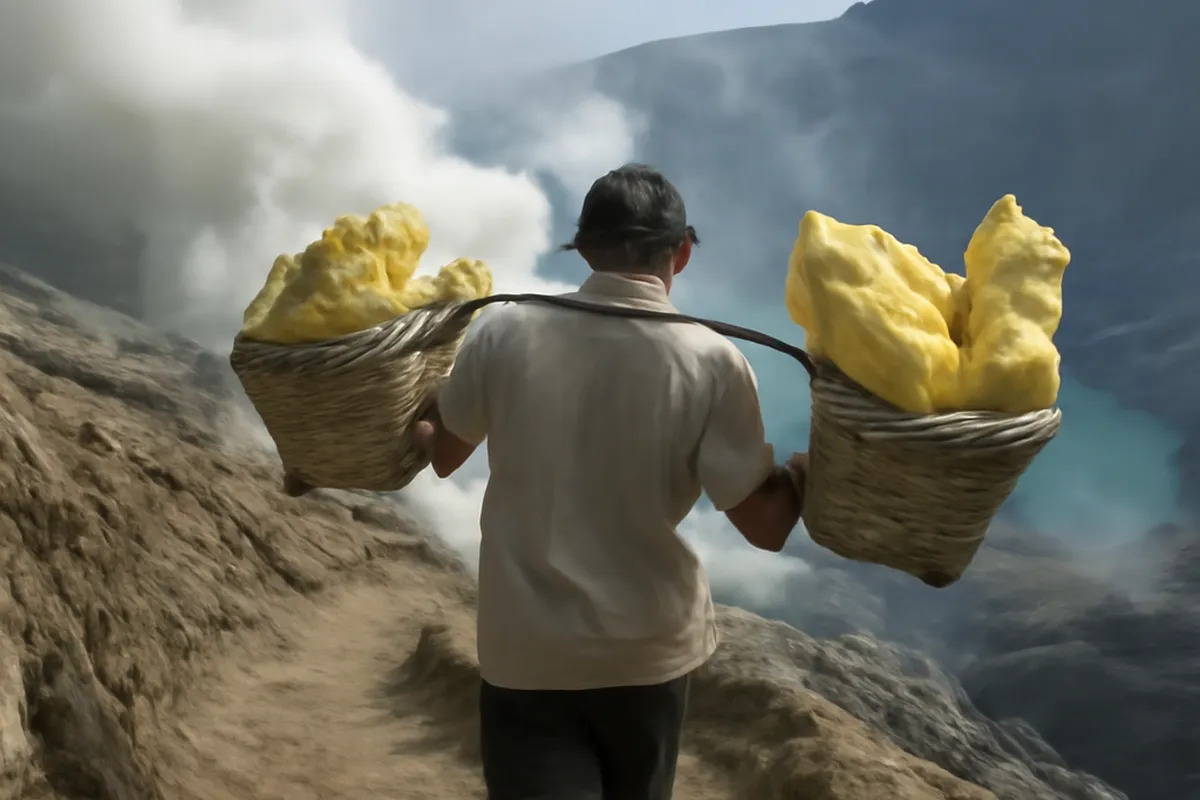 Porteur de soufre gravissant un sentier du volcan Kawah Ijen avec des paniers en osier chargés sur les épaules, fumerolles en arrière-plan